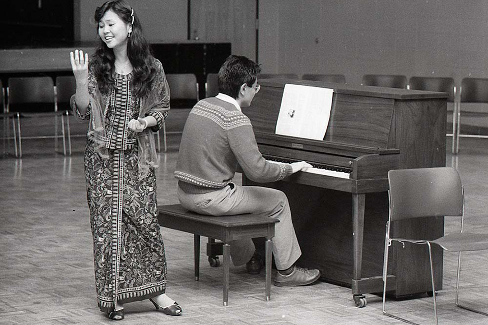 A woman stands by a piano while a man plays.