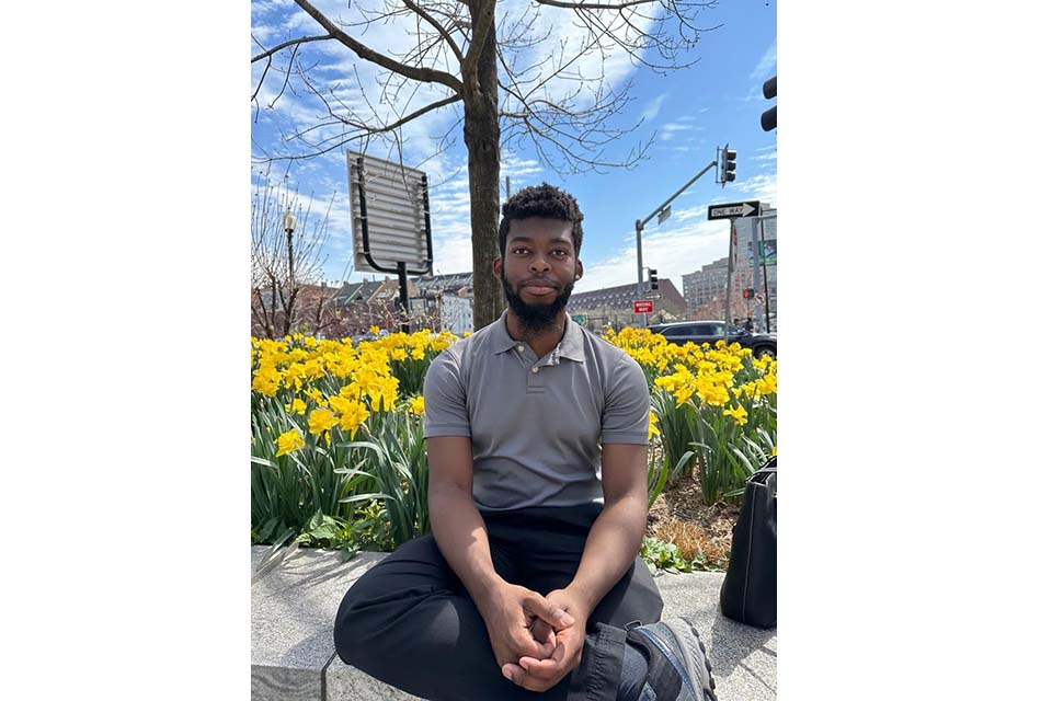 Abichael Belizaire sits on a stone wall in front of flowers in a city setting.