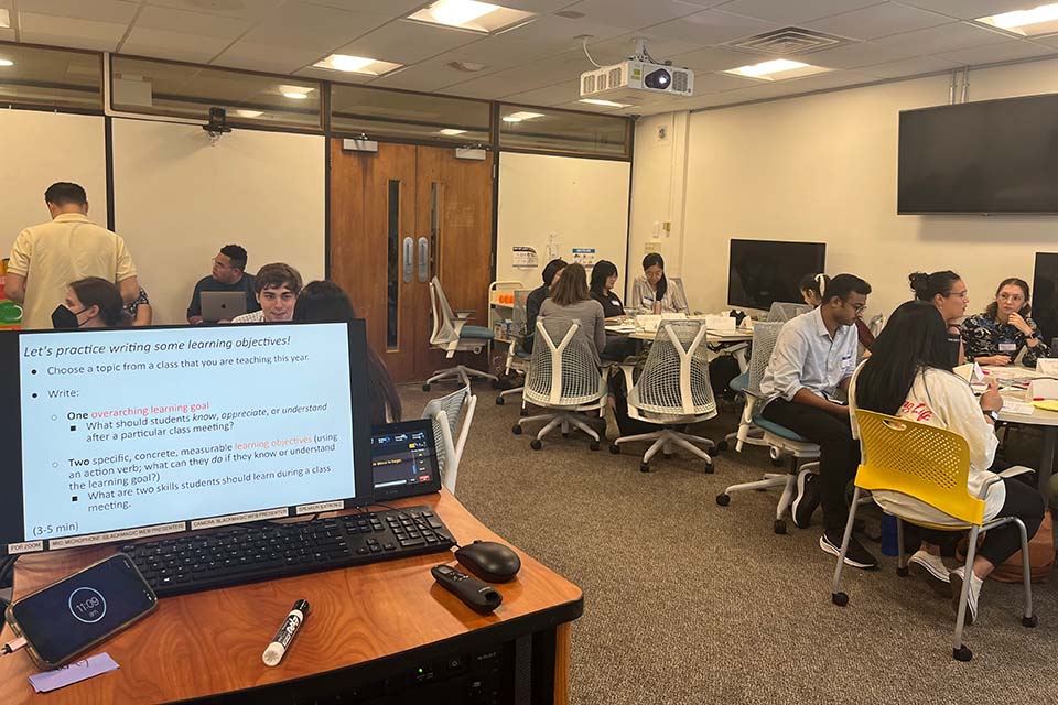 Students sit in small groups around tables. Text on a computer screen reads, "Let's practice writing some learning objectives!" and gives instructions: "Choose a topic from a class that you are teaching this year.  Write:  One overarching learning goal What should students know, appreciate, or understand after a particular class meeting?  Two specific, concrete, measurable learning objectives (using an action verb; what can they do if they know or understand the learning goal?) What are two skills students should learn during a class meeting." 