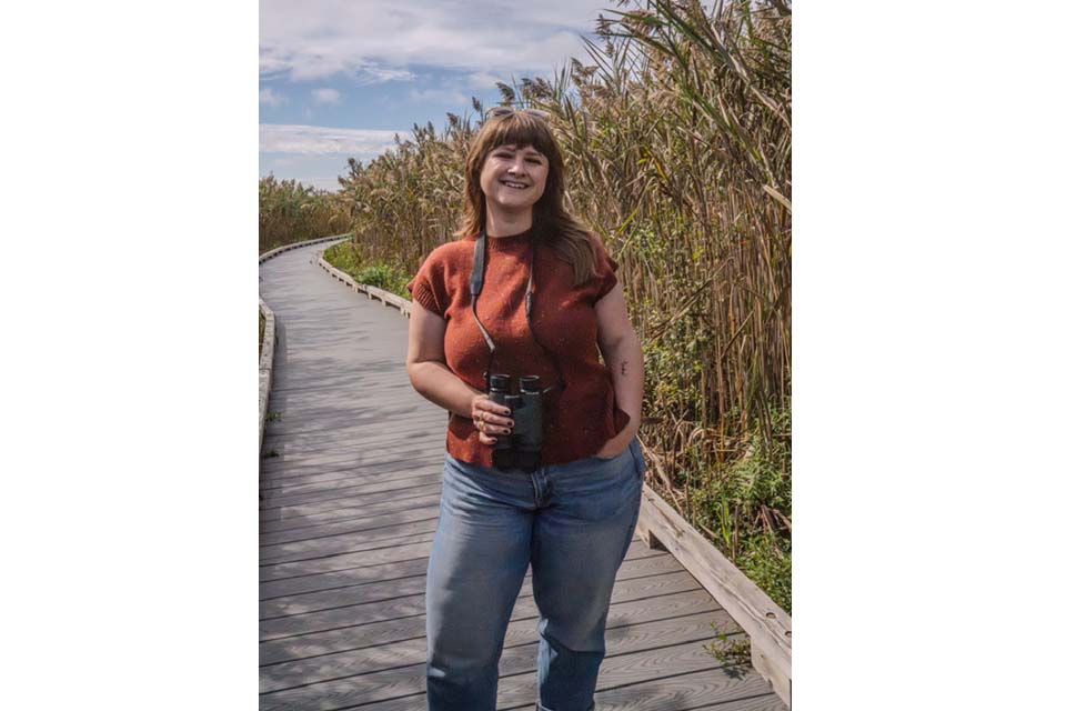 Jessi Brewer stands on a wooden walkway holding binoculars, next to some plants.
