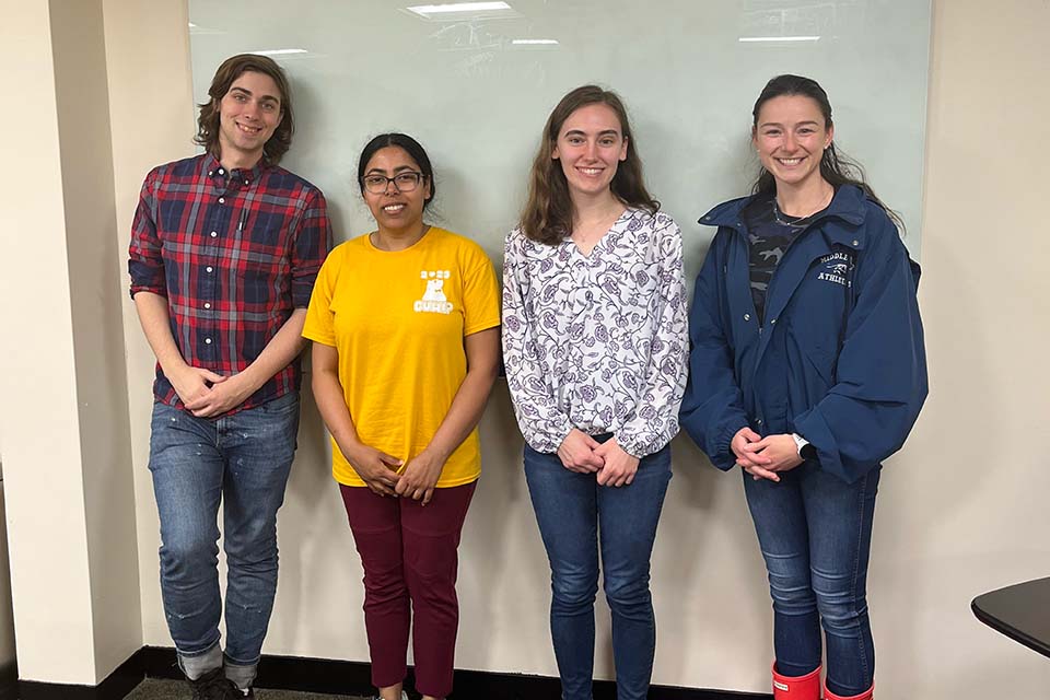 Nathanial Walker, Kanaya Malakar, Anne Silveira, and Emma McGuirk stand in a row in front of a whiteboard, smiling.