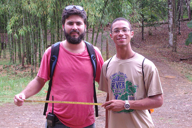 A Brandeis student stands with a local supervisor holding a tape measure in front of trees