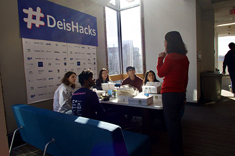 Students in a group sit around a table and listen to a mentor. Behind then hangs a large blue banner that reads "DeisHacks".