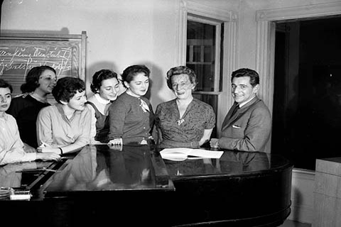 A black and white photo of Leonard Bernstein and Lillian Hellman standing with students around a piano.