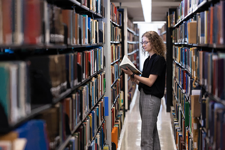 A student stands between bookshelves in the library reading a book