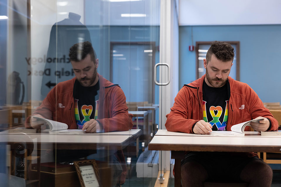 A student seated at a table looks down at a book. The window next to him displays his reflection.