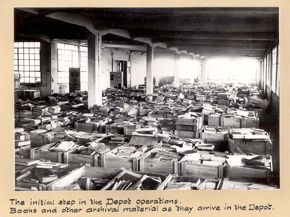 Black and white photograph of a large room filled with stacks of books. A handwritten note reads: The initial step in the Depot operations. Books and other archival material as they arrive in the Depot.