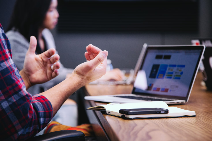 Person talking with their hands, sitting at a conference table with a laptop in front of them