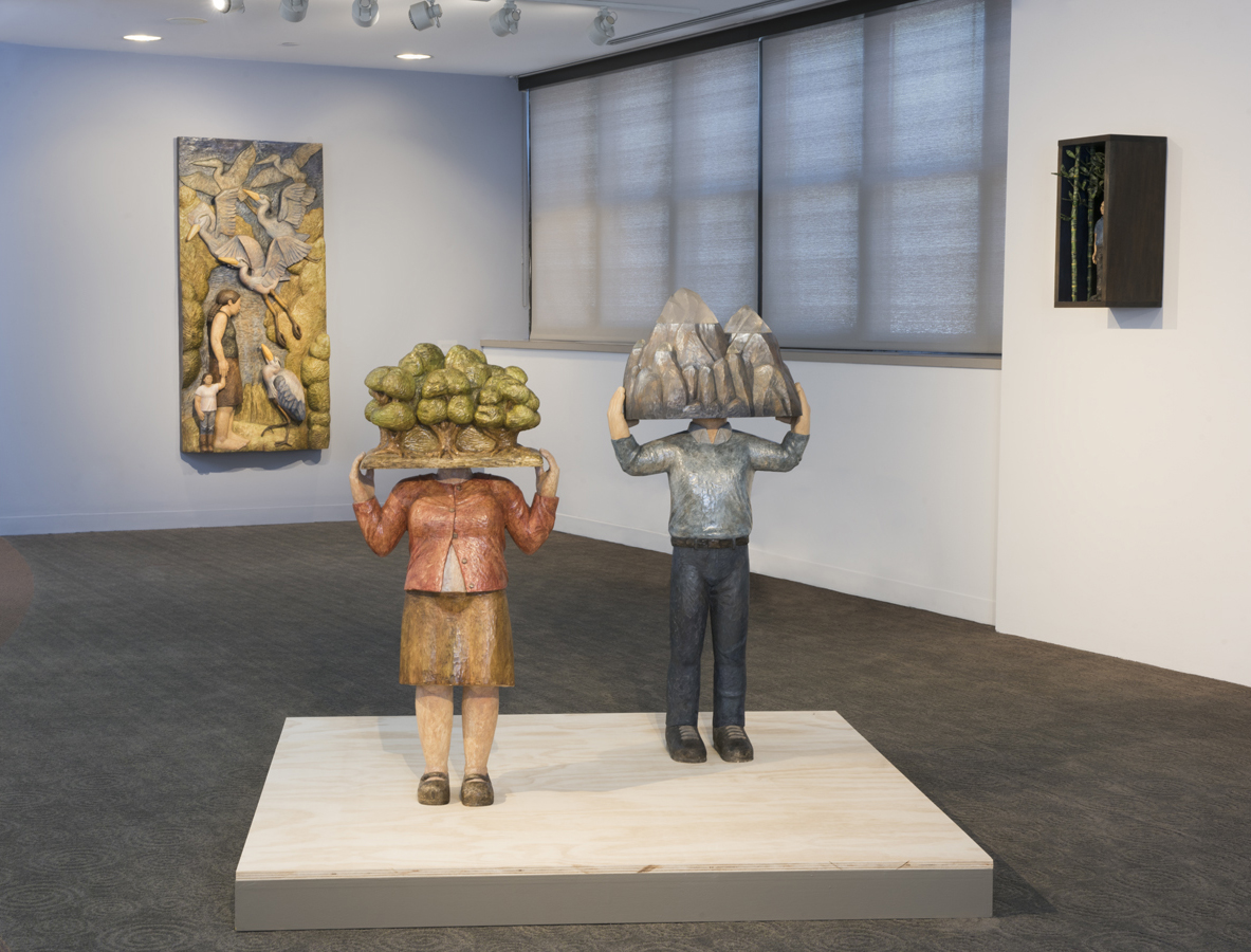 View of Kniznick Gallery installation of Sachiko Akiyama's exhibit "Long Hand Poem." Photo shows the scultpure “Origins,” 2014, wood, resin, paint, 46” x 19-½” x 11”.  It is displayed on a platform with 2 wooden figures.  Each figure has something from nature instead of a head. The woman has a forest.  The man has a mountain.  Both figures' hands are holding these natural forms in place. Two other artworks are on the walls behind the sculptures.  On the left is “On Finding Home,” a carved image made from polychromed wood and gold leaf, 40” x 8-½” x 9-½”. The other is a construction in a shadow box containing a figure and trees; only partially visible.