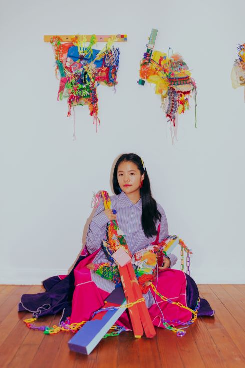 A colorful photograph of artist Loretta Park seated on the floor of her studio, holding work, with other work mounted above her on a wall. 